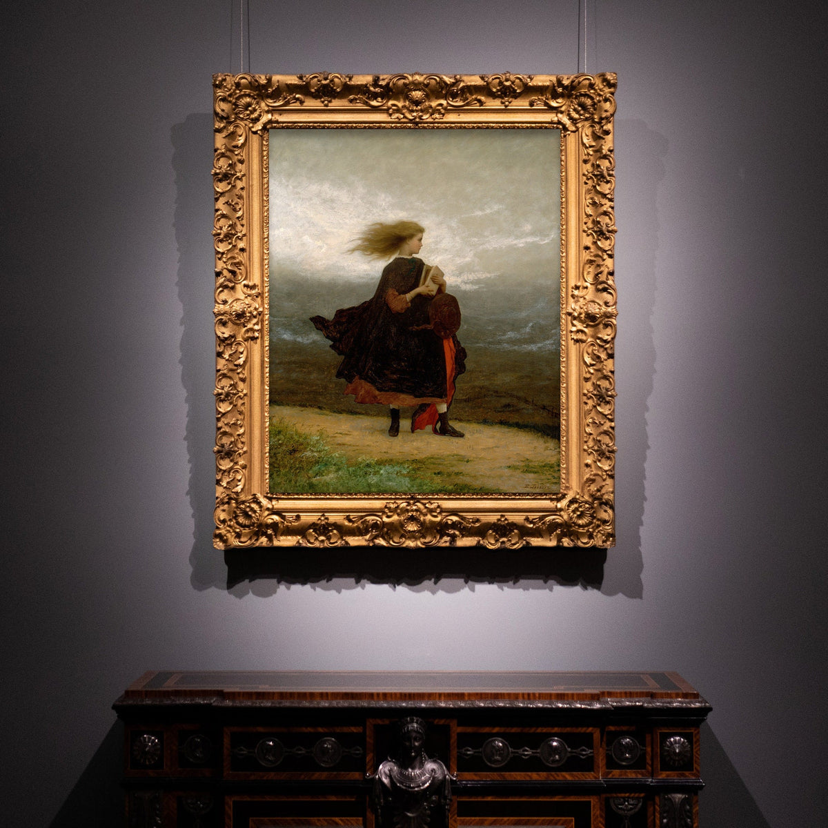 A fine art print in a very ornate gold frame hangs on a dark coloured wall. The picture depicts a windswept young lady walking across a landscape in a velvet coat and holding a book.