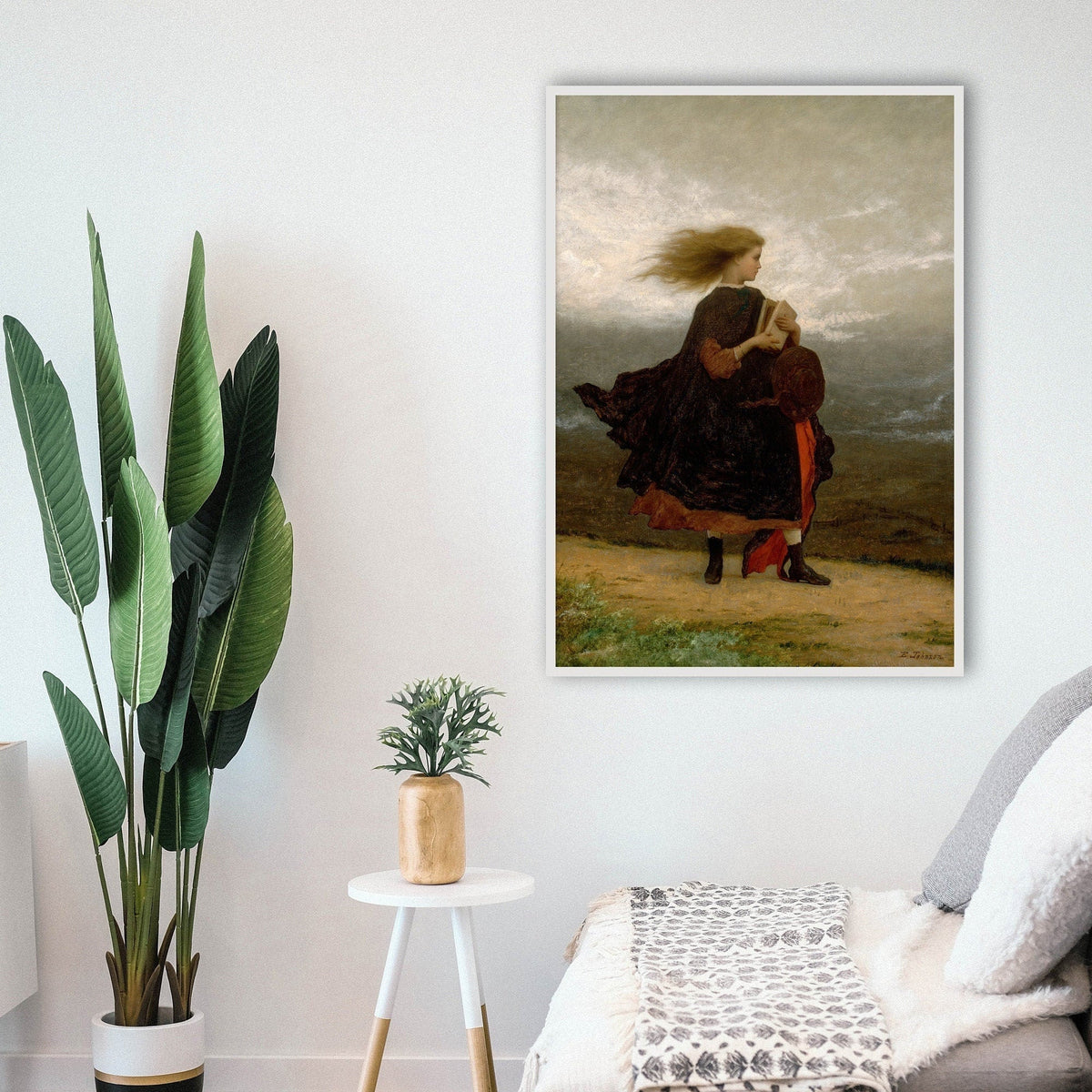 A fine art print in a living room setting with a sofa and plants. The picture depicts a windswept young lady walking across a landscape in a velvet coat and holding a book, her hair flowing in the wind.