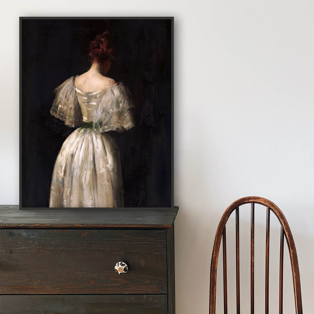 Wooden chair in front of a dark dresser with a print of a woman in a long dress resting against the wall.