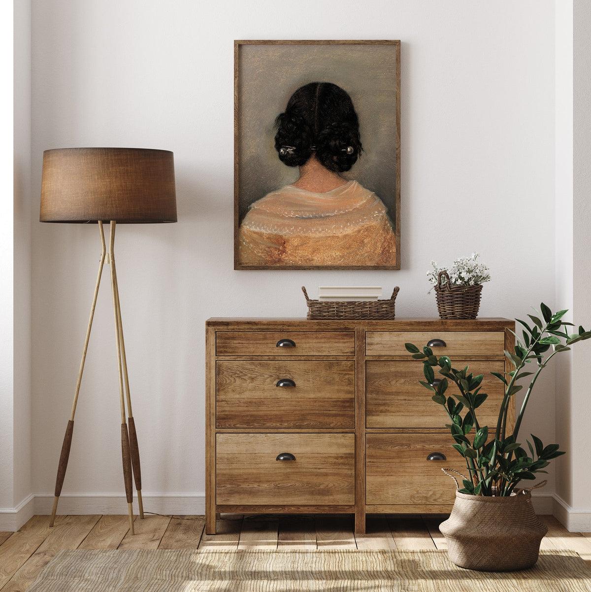 Wooden dresser with a lamp, plant, and framed print of a woman from behind hanging on a white wall.