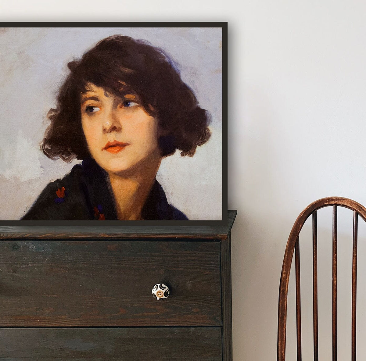 A fine art print of a portrait in oil of a young lady from the Art Deco period. The print is displayed in a thin black frame on top of a chest of drawers.
