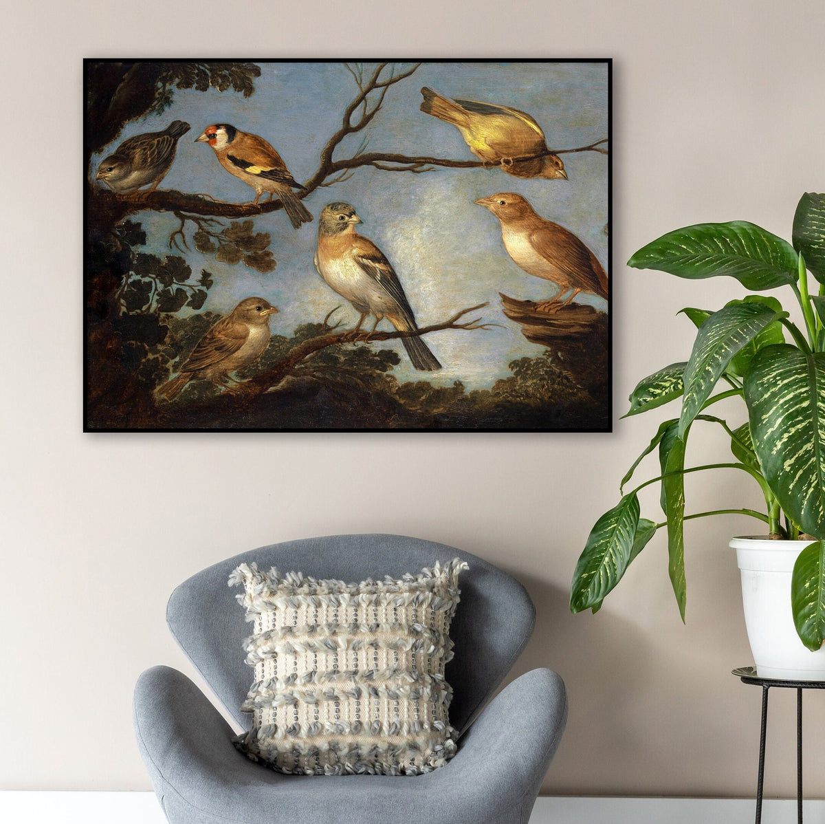 A framed print of garden birds gathered together on the branches of a tree. The print is displayed hanging on the wall in a thin black picture frame above a grey-blue chair.