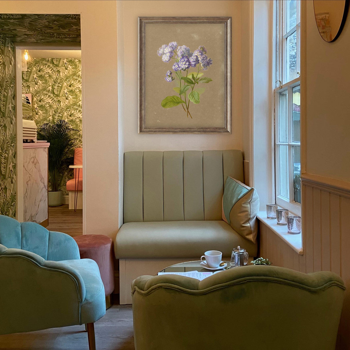 A fine art print of a delicate botanical artwork that features a sprig of purple heliotrope. The print is displayed hanging on a wall by a window in a wooden picture frame in a green-themed living room.