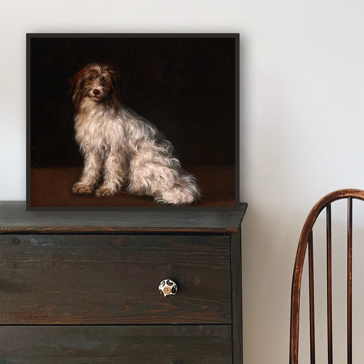 Wooden chair and black dresser with a framed picture of a dog on a dark background. The print is resting against a white wall.