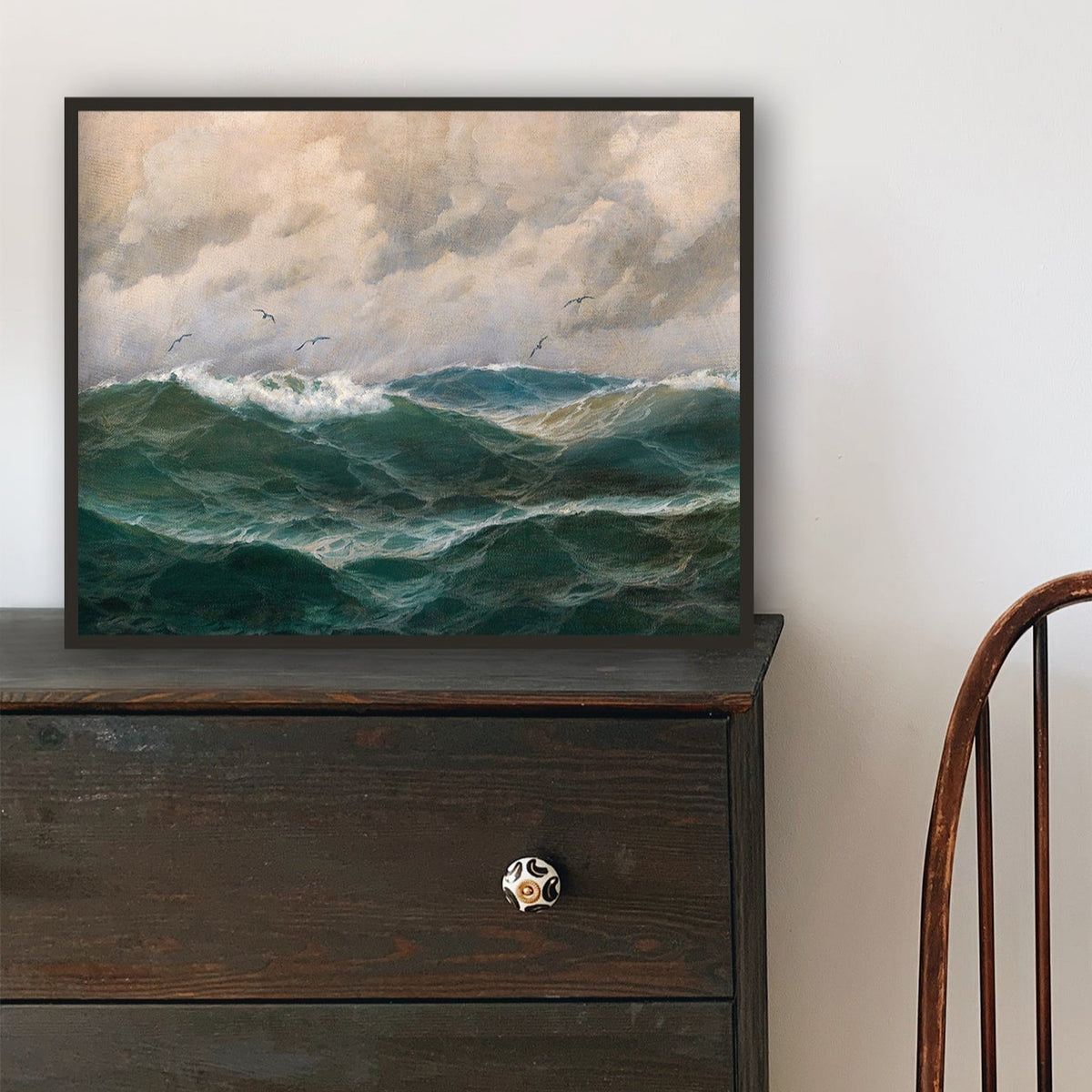 Wooden chair in front of a black dresser with a framed seascape on top