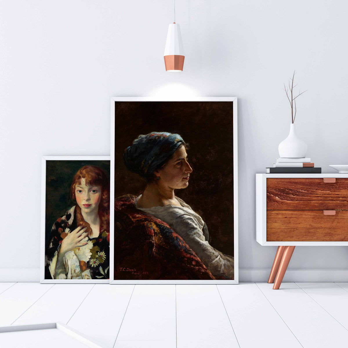 Two fine art prints of classical portrait paintings sit on the floor against a white wall with a modern wooden side table.