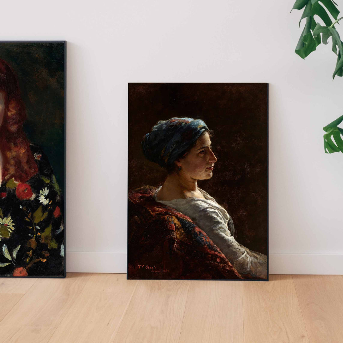 Two fine art prints of oil painting portraits sit on the floor, resting against a white wall with a plant in the corner.