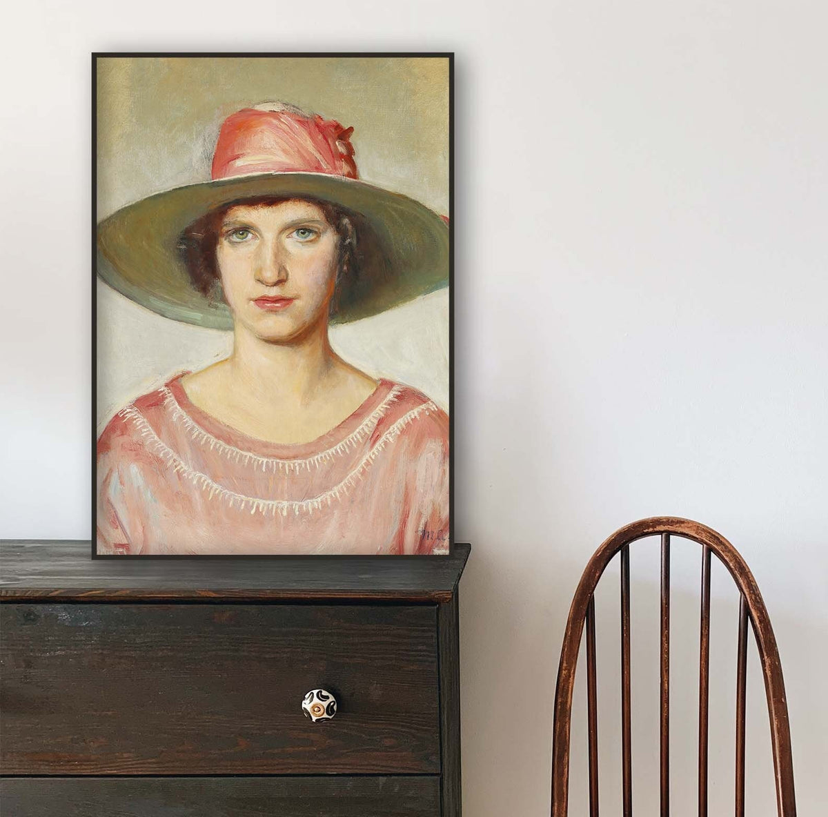 A fine art print of an Art Deco oil painting of a portrait of a lady. The print is displayed in a thin black frame resting on top of a dark wood chest of drawers.
