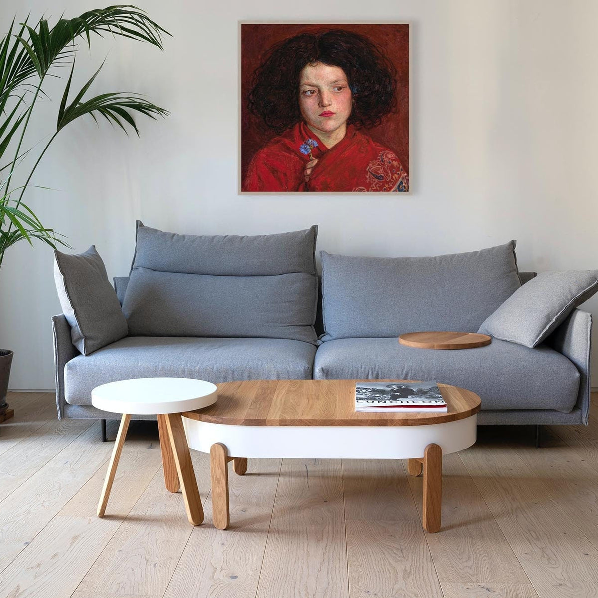 A fine art print of a beautiful oil painting of a young girl in a red shawl from mid 19th century. The print is displayed in a thin picture frame above a large grey sofa.