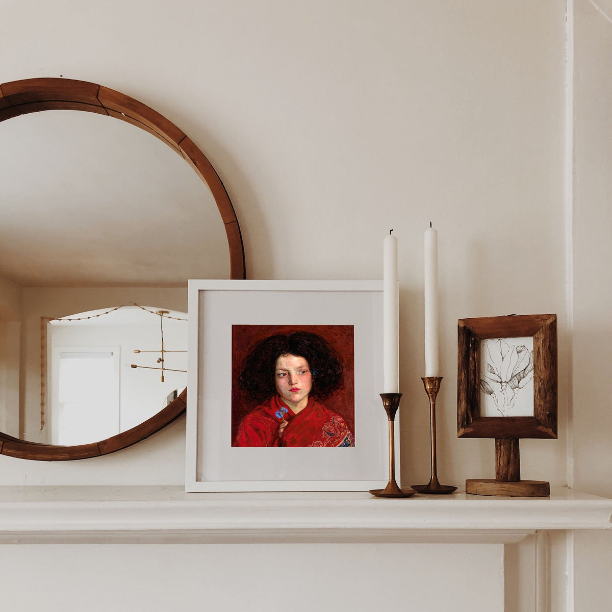 A fine art print of a beautiful oil painting of a girl in a red shawl from the mid 19th century. The print is displayed mounted in a white picture frame on top of a white mantlepiece.