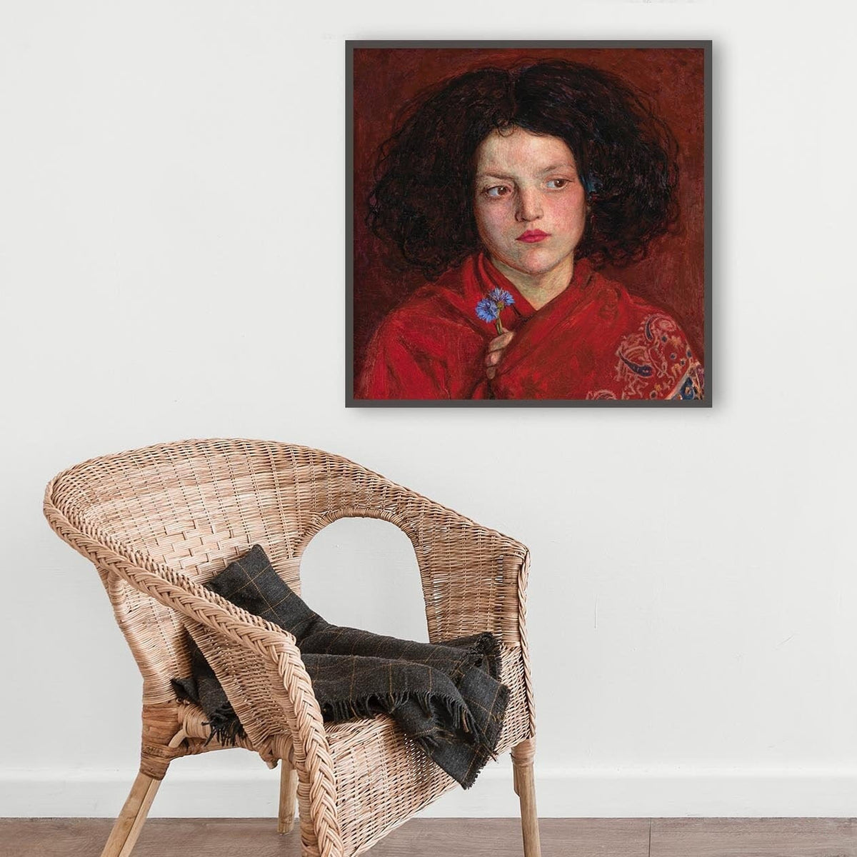 A fine art print of a beautiful oil painting of a young girl in a red shawl from mid 19th century. The print is displayed hanging on the wall in a dark picture frame above a wicker chair.