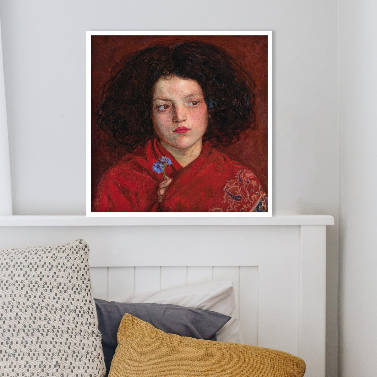 A fine art print of a beautiful oil painting of a young girl in a red shawl from mid 19th century. The print is displayed in a thin white picture frame resting on top of a white headboard.