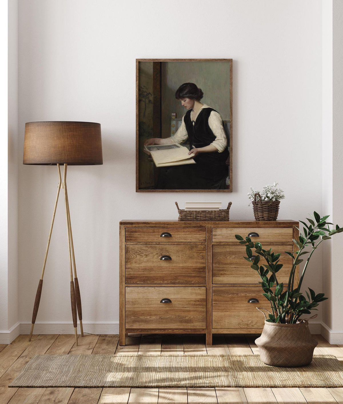 A wooden dresser with a lamp, plant, and framed print of a woman reading, in a room.