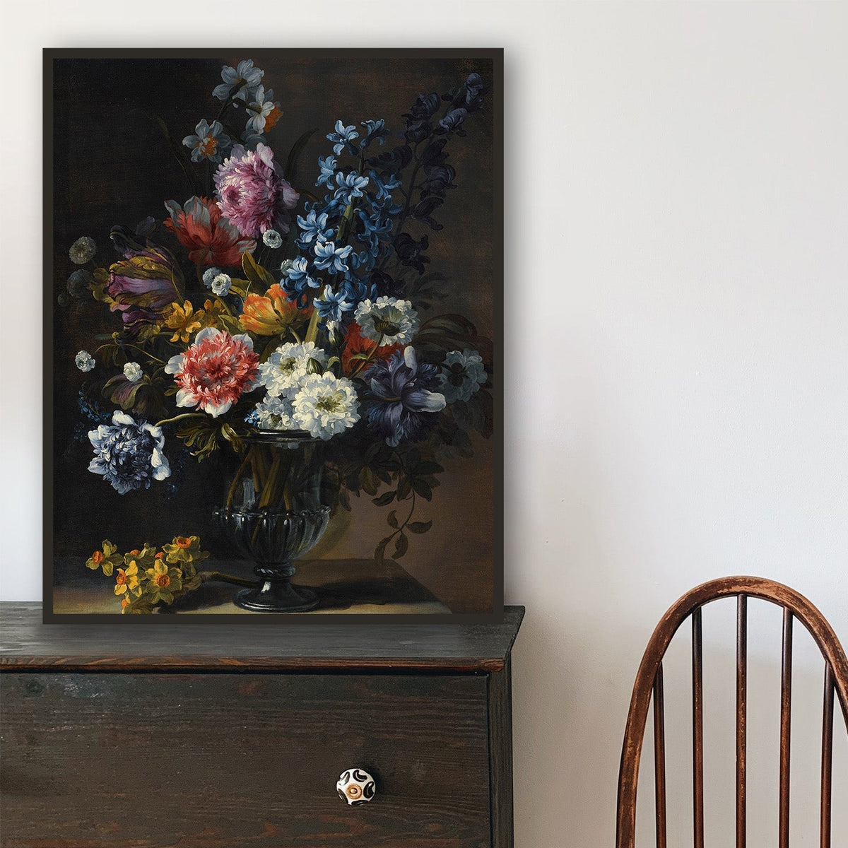Wooden chair in front of a black dresser with a floral painting on a white wall.