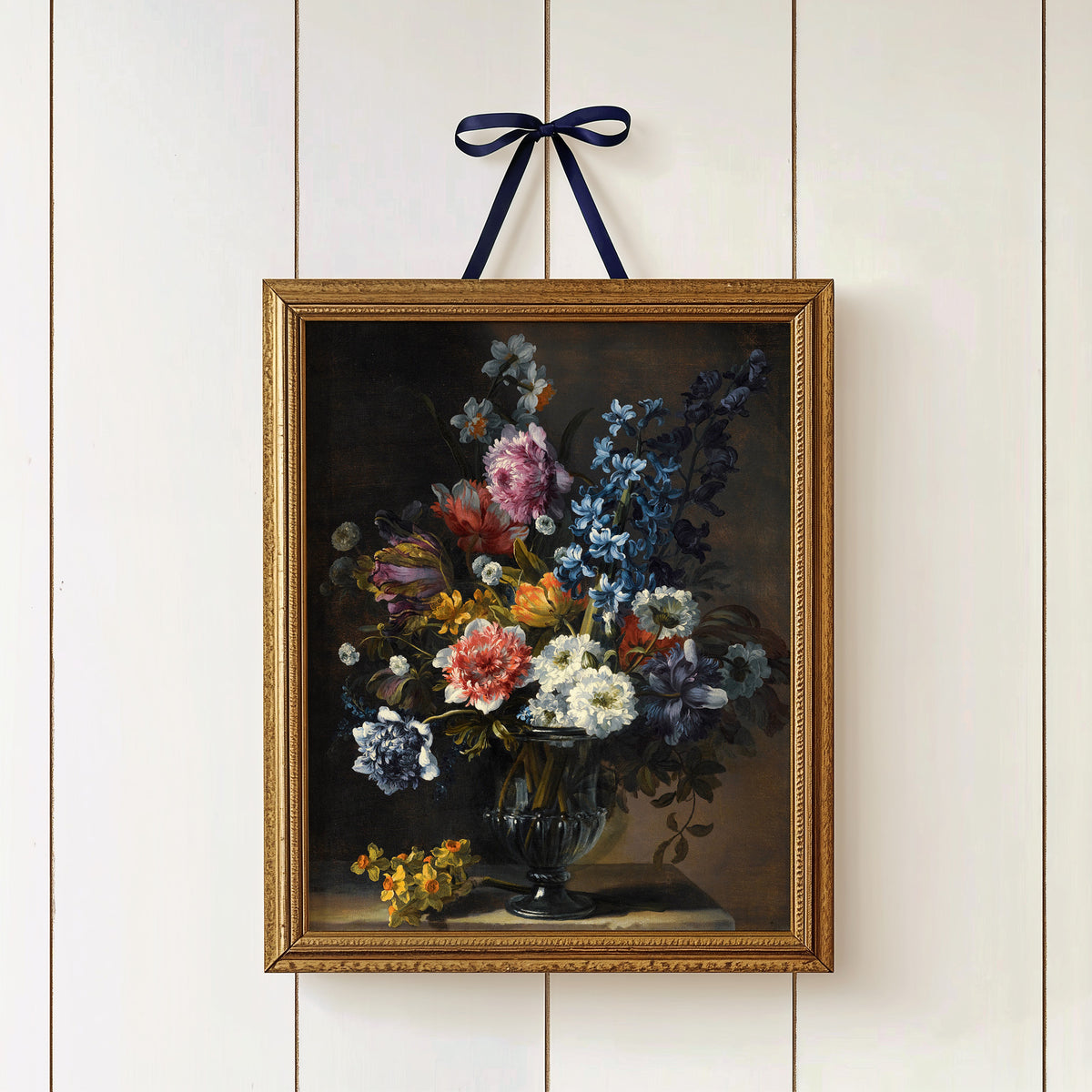 Gold-framed print of a floral arrangement on a white paneled wall, hung with a ribbon.
