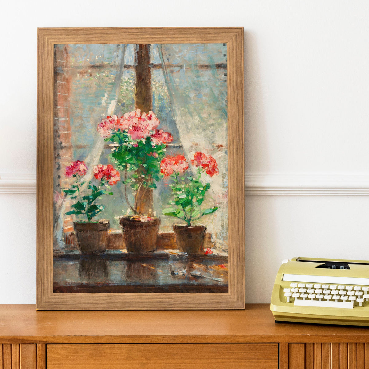 A picture of red and pink geraniums in terracotta pots sat on a windowsill, is framed in a light wood picture frame, and sits on top of a cabinet next to a typewriter.