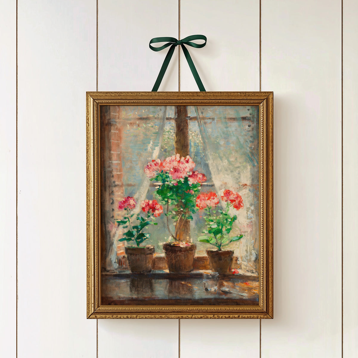 Framed print of potted flowers hanging on a white paneled wall in a gold frame with a green ribbon.