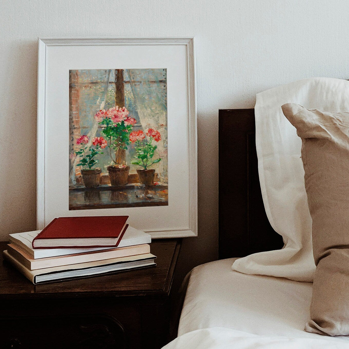 A print of red and pink geraniums in terracotta pots sat on a windowsill, framed in a white picture frame on a bedside table.