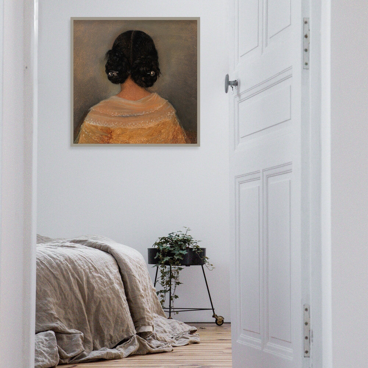 Framed artwork of a woman from behind with dark hair on a light gray background hung in a thin frame on a white wall above a bed and plant.