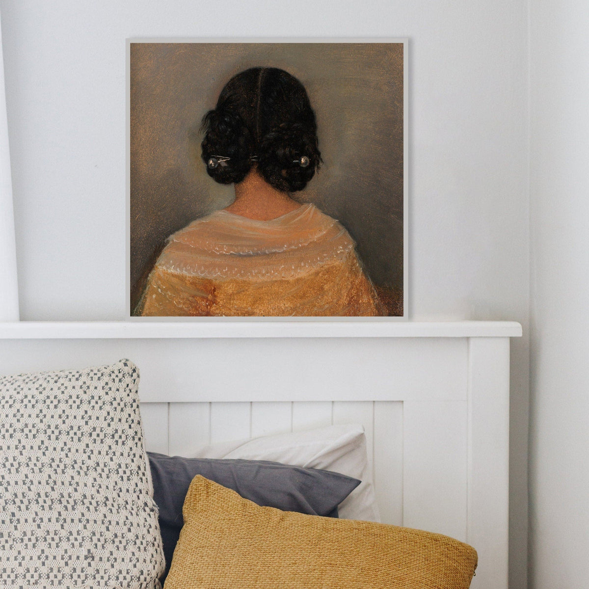 Framed artwork of a woman from behind with dark hair on a light grey background in a white frame resting on top of a bed with cushions.