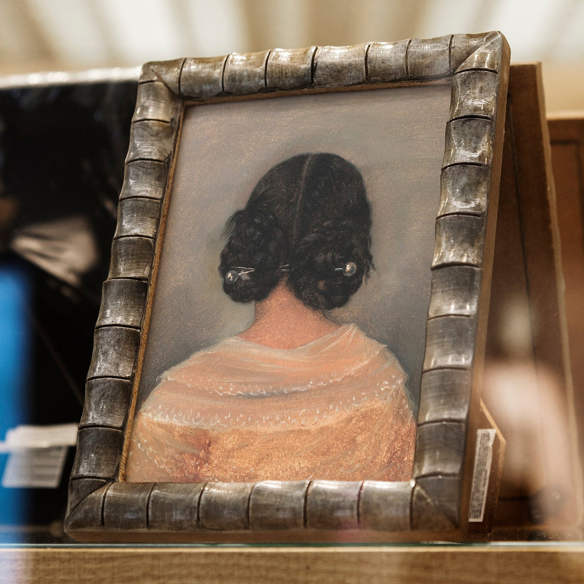 Small portrait of a lady from behind with dark hair in an ornate frame on a blurred background.