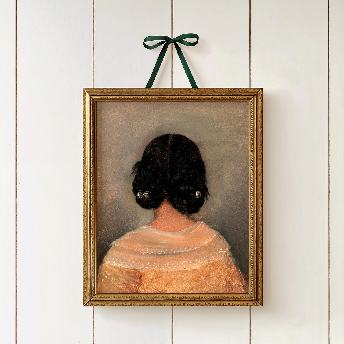 Framed print of a woman from the back with dark hair and a lace top, hanging on a white wall in a gold picture frame and green ribbon.