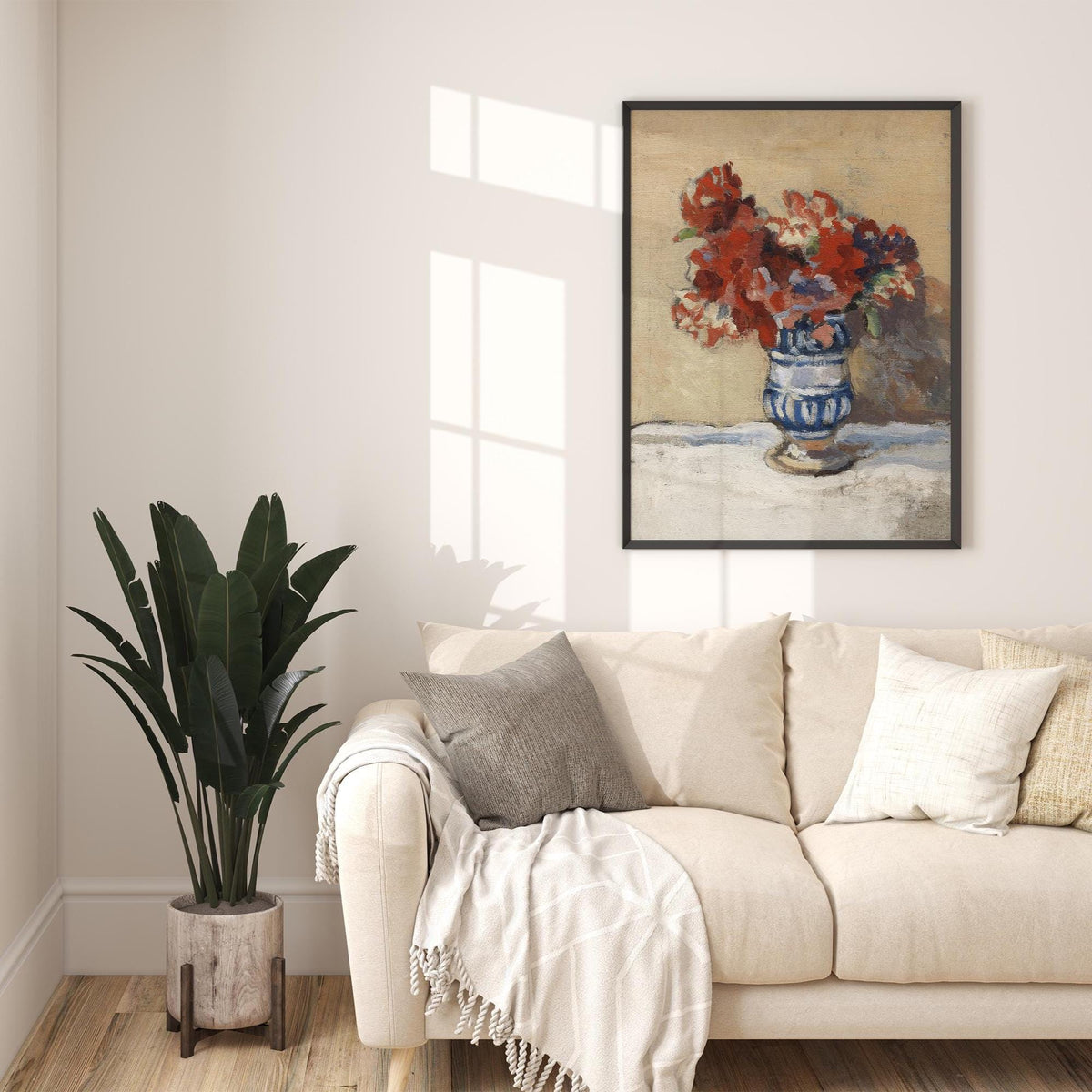 A fine art print of a still-life oil painting of red flowers in a blue and white striped vase. The print is displayed in a thin black picture frame above a cream sofa in a comfy living room.
