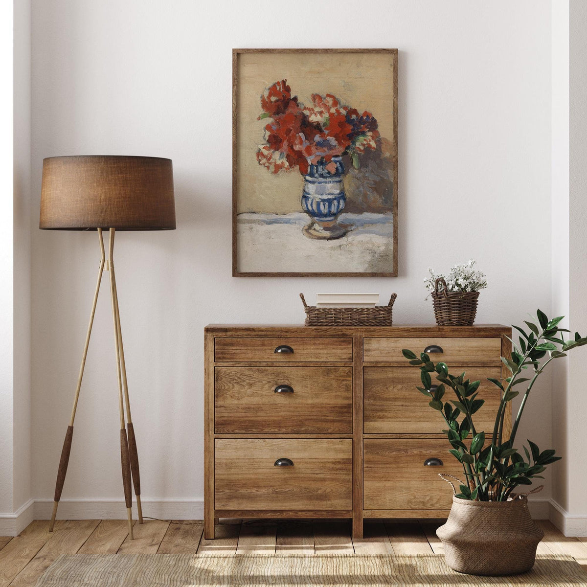 A fine art print of a still-life oil painting of red flowers in a blue and white striped vase. The print is displayed hanging on the wall in a thin wooden picture frame above a wooden chest of drawers in a modern living room.