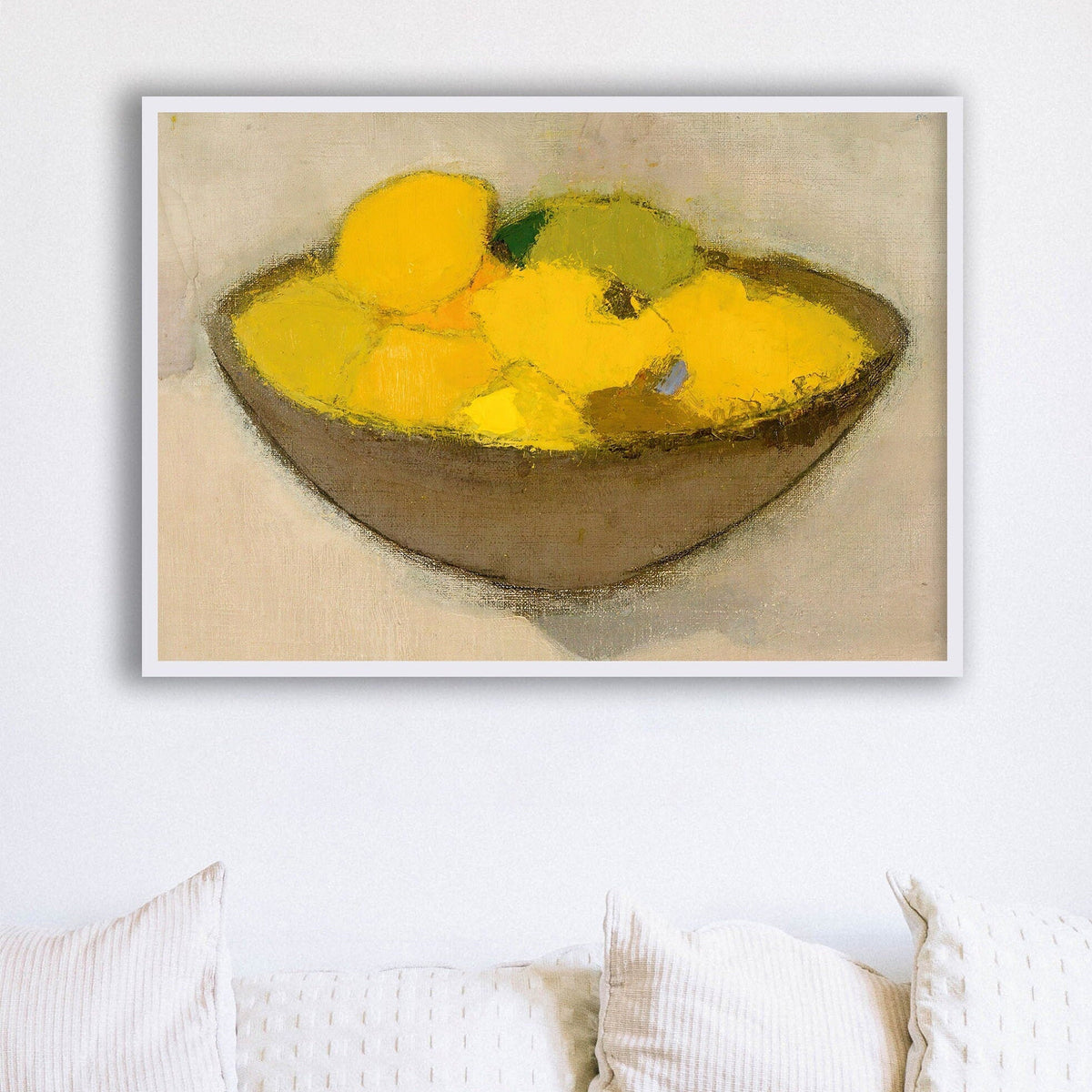 Framed print of a bowl with yellow lemons on a neutral background, hung above some white cushions.