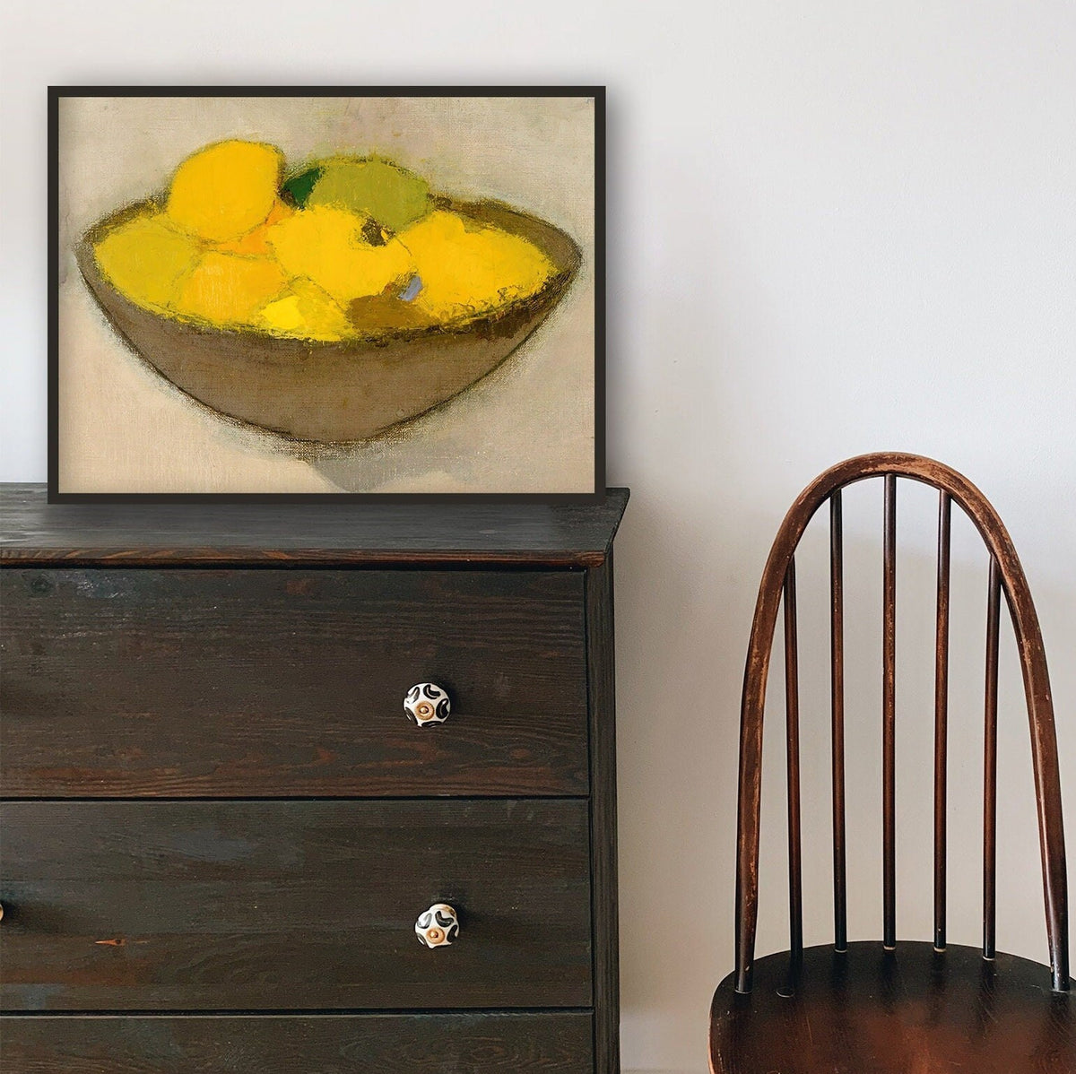 Framed artwork of a bowl of lemons on a wooden dresser next to a chair.