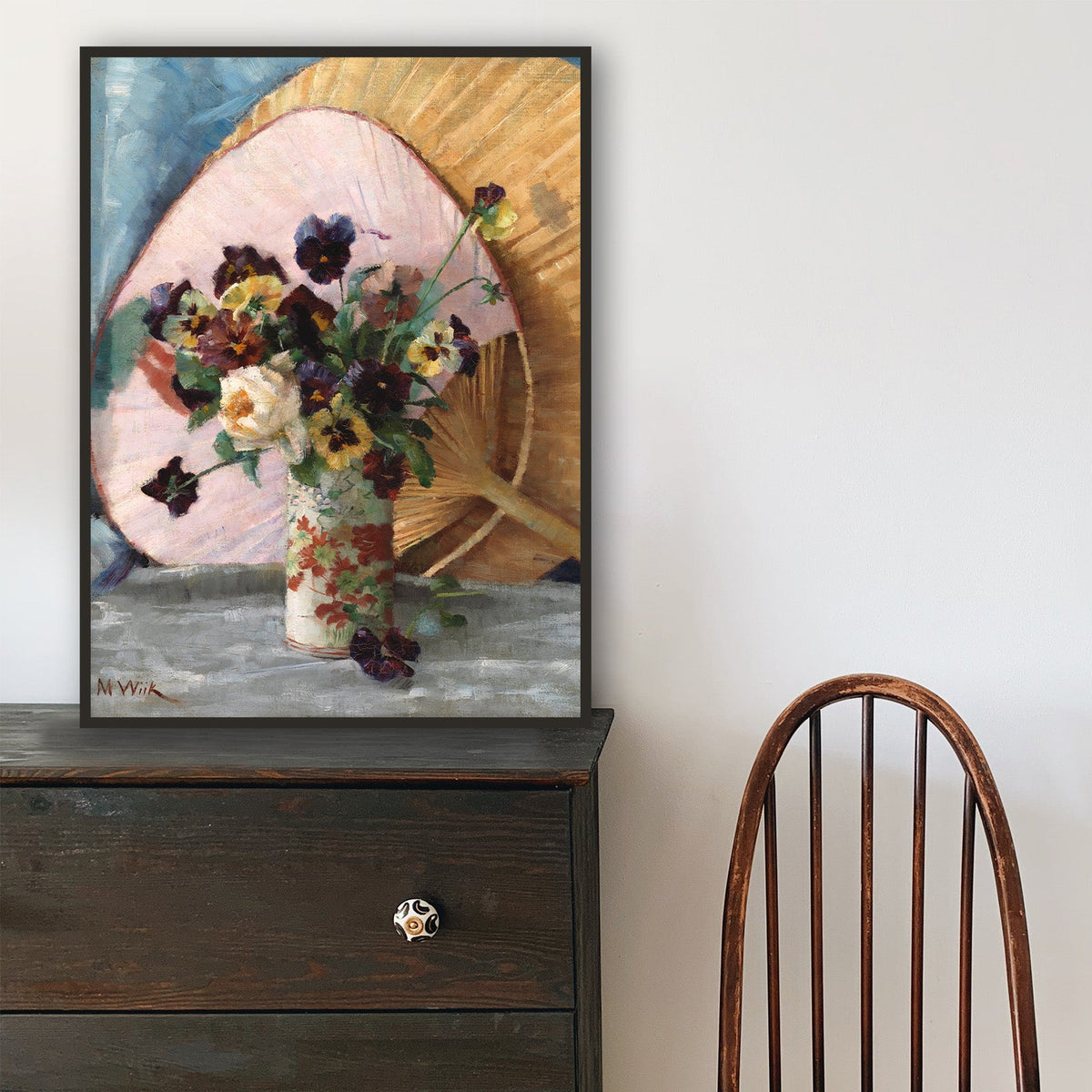 Wooden chair in front of a black dresser with a framed fine art print of a floral still-life on top.
