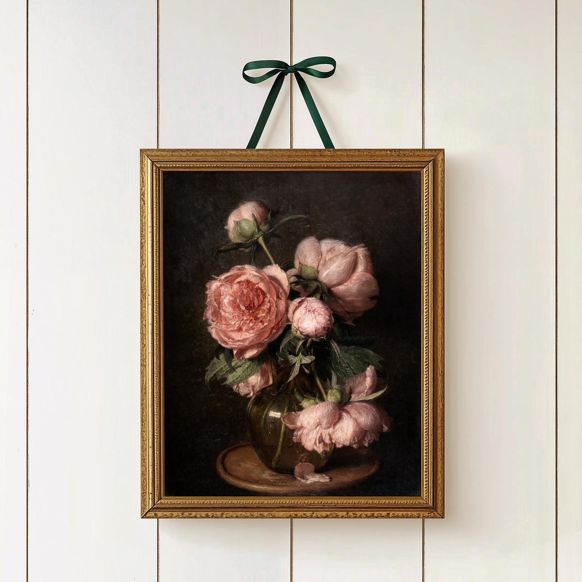 A gold framed fine art print of pink peonies on a dark background, hung on a white wall with a green ribbon