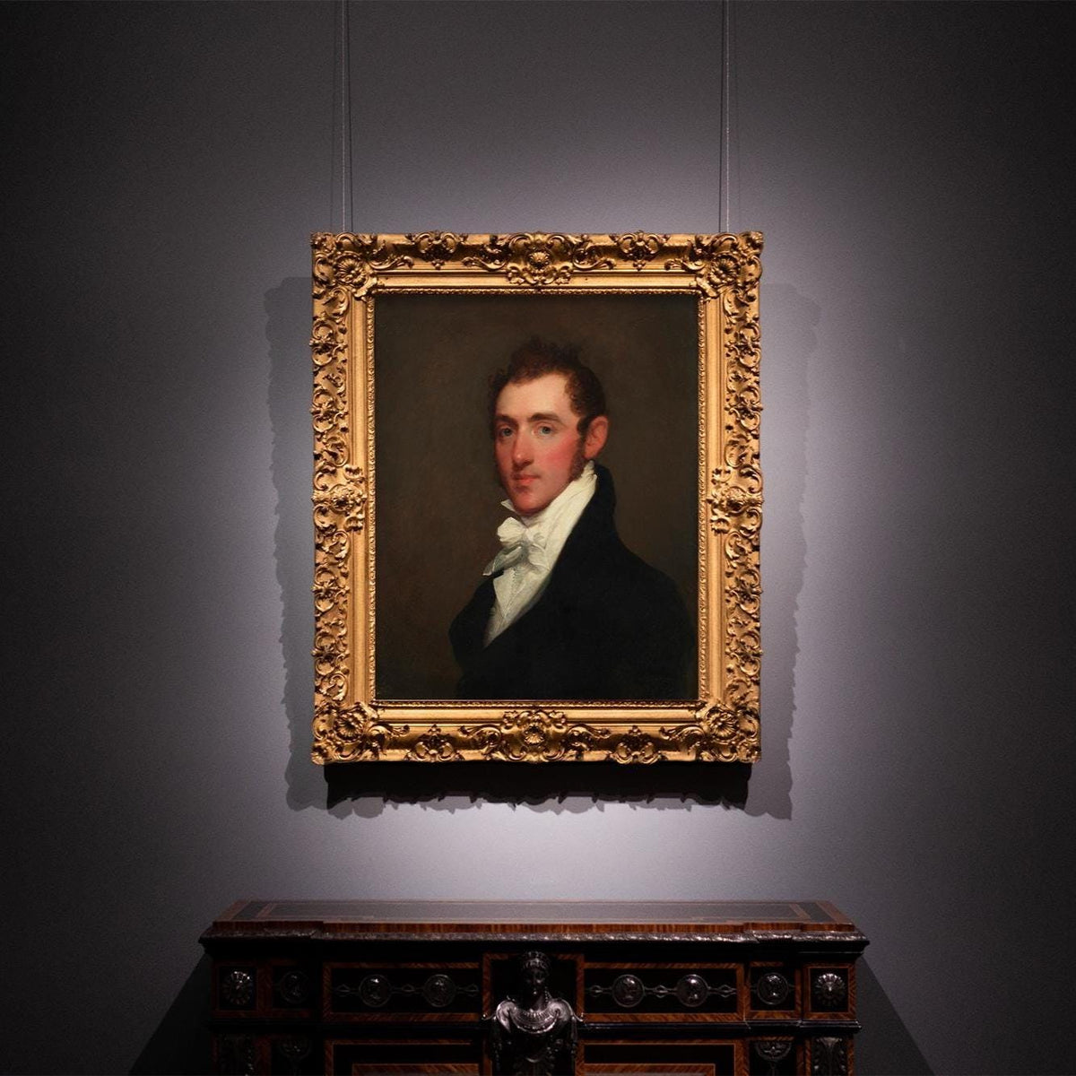 A fine art print of a Regency-era gentleman. The print is displayed in an ornate gold frame on a dark painted wall.