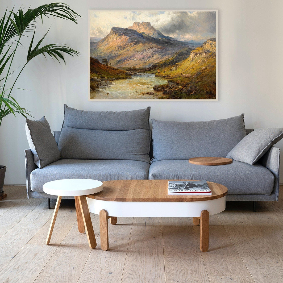 A fine art print of an oil painting depicting dramatic scenery and beautiful sunlight hitting the hills around Ben Lomond in Scotland. The print is displayed above a large grey sofa in a living room.