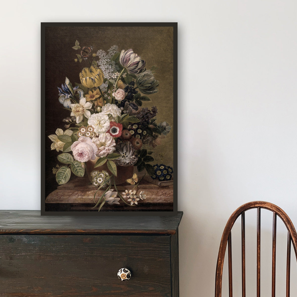 Framed floral print on a wooden dresser with a chair to the side.